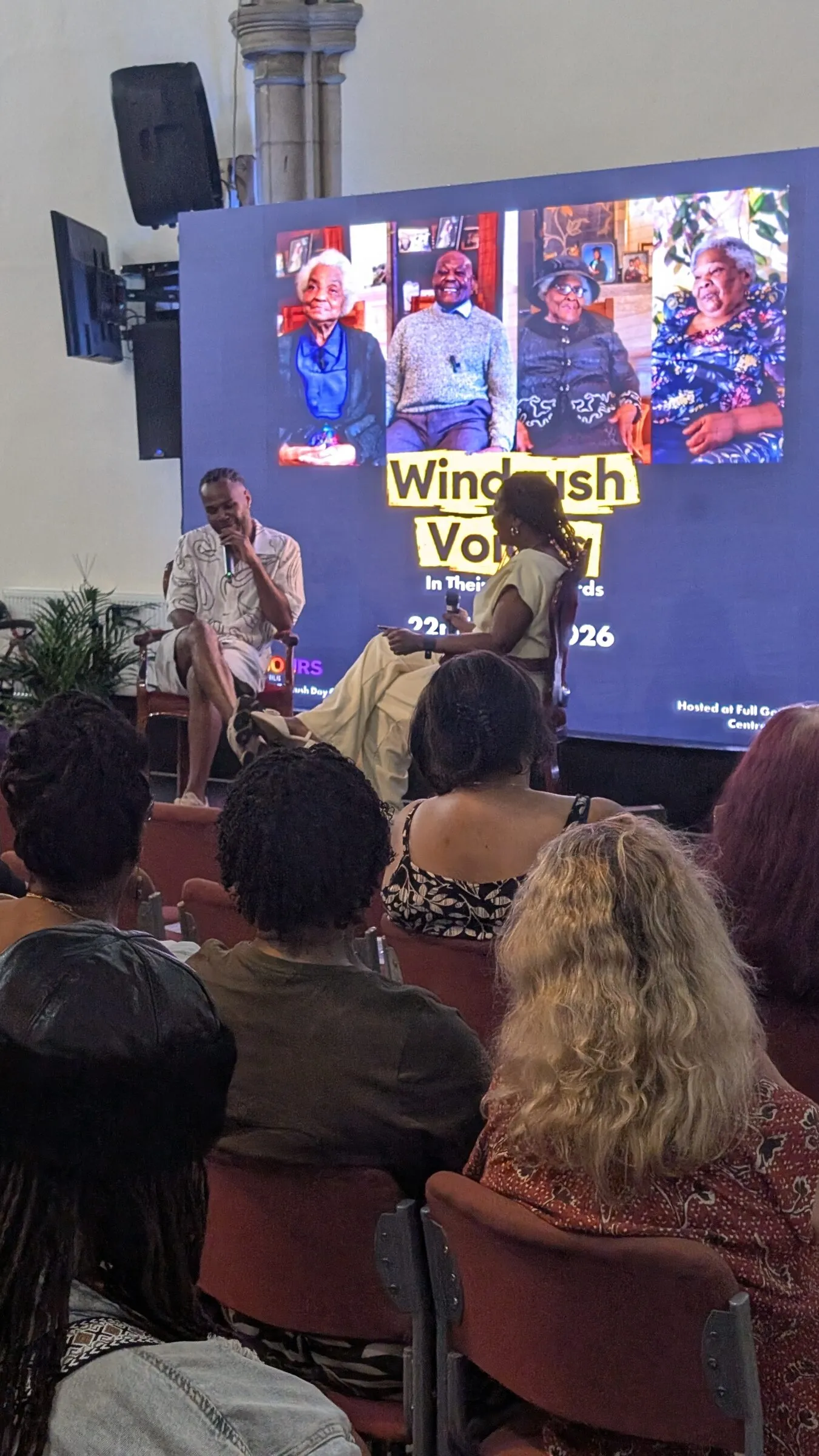 A conversation on stage at Windrush Voices, with the event title on the screen behind and the audience in the foreground.